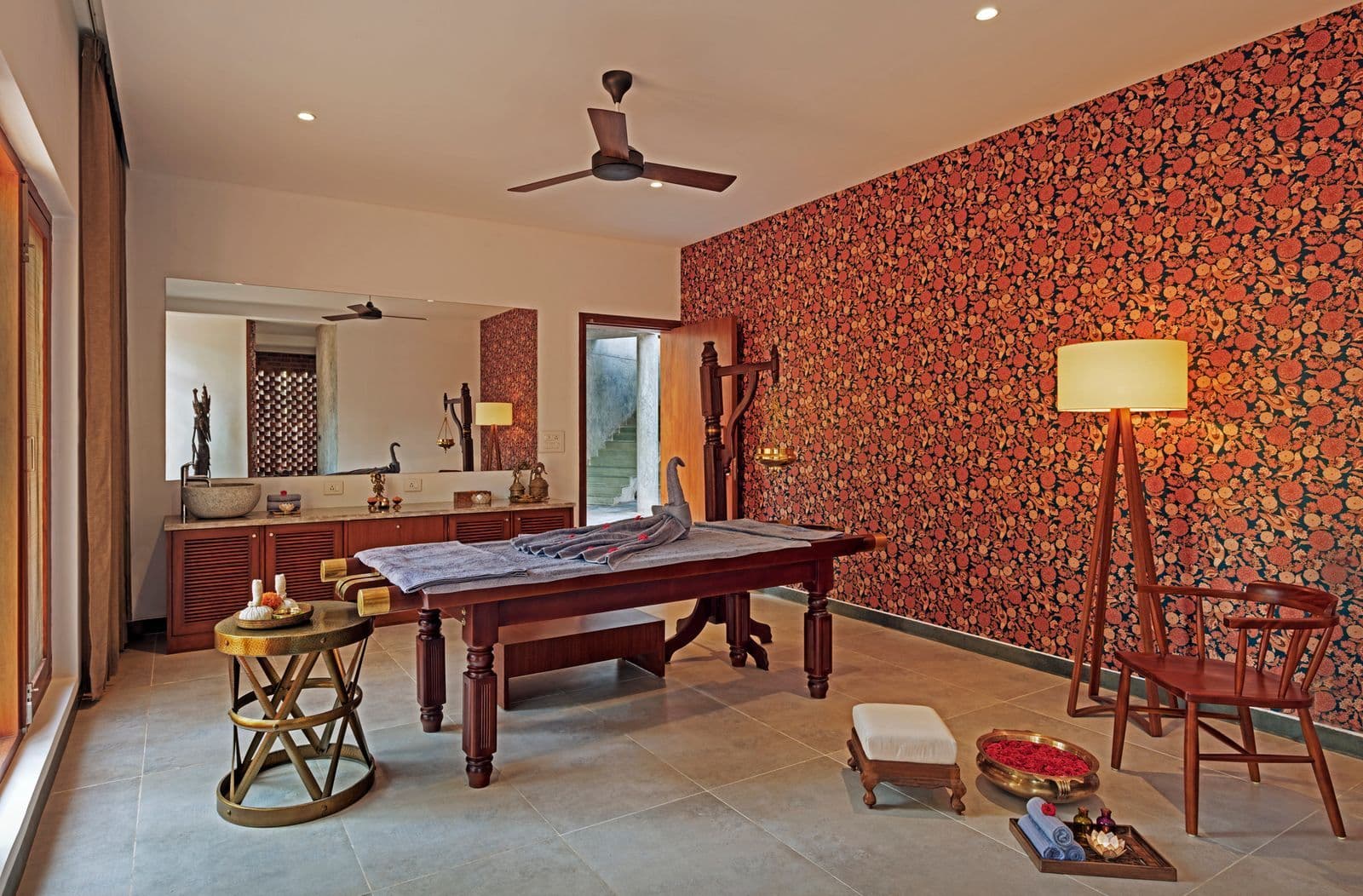 An Ayurvedic treatment room with a carved wooden massage table set against a richly patterned floral accent wall